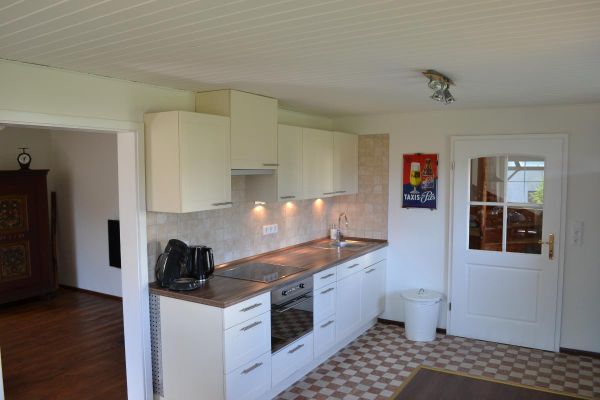 Eine weiße Küchenzeile mit Holzarbeitsplatte steht auf einem kariertem Fliesenboden neben einer weißen Zimmertür mit Sprossenfenster; im Durchgang zum Wohnzimmer ist ein Bauernschrank sichtbar.