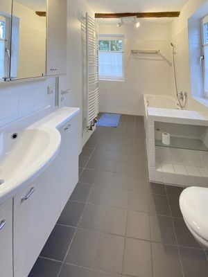 Badezimmer mit Badewanne, Waschbecken, Heizkörper und Fenster, helle Fliesen und grauer Boden.