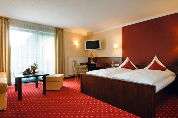 Großzügiges Schlafzimmer auf rotem Teppichboden mit weißen Punkten. Doppelbett und Rückwand aus dunklem Holz. Bettrückwand deckenhoch rot gestrichen. TV-Gerät über dem Schreibtisch. Gegenüber Sofa, Tisch und Hocker. Bodenlange Gardinen und Vorhänge vor der Fensterfront.