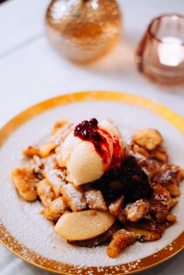 Auf dem Bild ist ein Teller (Goldrand) mit Kaiserschmarren, Früchten und Eis auf einem Tisch stehend zu sehen. Im Hintergrund ist eine Kerze zu sehen.