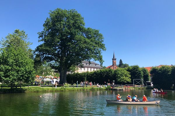 Eine großzügige Parkanlage mit Teich und Schloss ist zu sehen. Es ist sommerlich warm und die Menschen flanieren. Auf dem Teich sind Kanufahrer unterwegs. Im Hintergrund ist ein roter Kirchturm zu sehen.