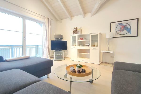 Neben einer großen Fensterfront und unter einem offenen, hellen Dachstuhl steht ein graues Ecksofa vor einem runden Glastisch auf Rollen. Zwischen einem Fernseher auf einer TV-Säule und einem Edelstahlbord, auf dem eine Lampe mit weißem Schirm steht, befindet sich ein weißer Wohnzimmerschrank mit Dekoartikeln. Ein buntes Bild hängt an der Wand.
