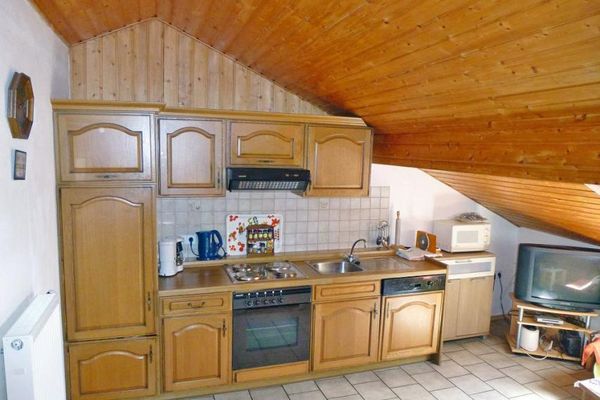 Im Raum mit holzvertäfelter Mansarde steht eine Holzküchenzeile. Daneben befinden sich eine Mikrowelle auf einer kleinen Kommode sowie ein Fernseher und weitere Geräte auf einem TV-Regal.