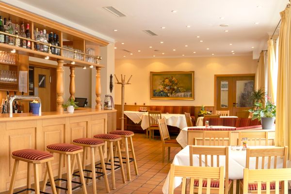 Das Bild zeigt ein gemütliches Restaurant mit einem Holztresen mit Barhockern davor, mehrere Tische mit weissen Tischdecken und Holzstühlen. Die Tische sind mit Blumendeko versehen, an der Wand hängt ein Blumenbild. Der Boden hat helle Fliesen.