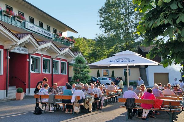Vor dem Hotel Gasthof Dilger laden Bierbänke und Tische unter einem großen Sonnenschirm zum Verweilen ein. Zahlreiche Gäste genießen die Bewirtung im Freien.