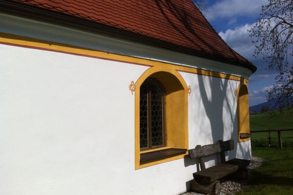Der Betrachter blickt auf eine Parkbank welche sich außen an einer Kapelle befindet und dem Pausierenden einen herrlichen Ausblick auf die Alpenkette bietet.