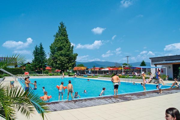Schwimmbecken mit Rutsche. Im und am Wasser tummeln sich die Badegäste. Liege- und Spielwiese mit Sonnensegel bieten Spaß und Erholung.