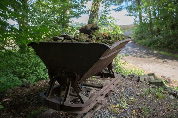 Eine eiserne Eisenlore steht gefüllt mit Gestein auf einem Schotterstück mitten im Wald an einem Weg.