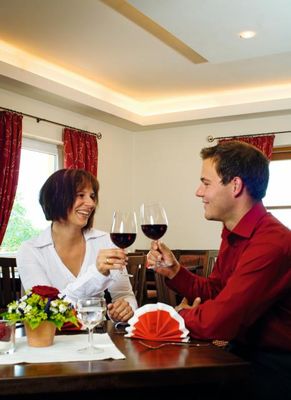 Ein Paar sitzt an einem gedeckten Tisch und prostet sich mit Rotwein zu. An den Fenstern hängen rotgemusterte Vorhänge.
