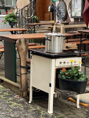 Blick auf ein Arrangement an einer Holzterrasse vor einem Gebäude, auf der zahlreiche Brauereigarnituren stehen. Im Vordergrund ist ein alter, weißer Ofen das zentrale Hauptmotiv. Die Ofenklappe ist geöffnet und ein Blumenarrangement in einen gusseisernen Bräter gepflanst befindet sich darauf. Auf dem Herd sthet ein Topf und eine Holzlaterne. Im Eingangsbereich des Fachwerkhauses im Hintergrund befinden sich mehr Alltagsgegenstände, die an einen Flohmarkt erinnern.