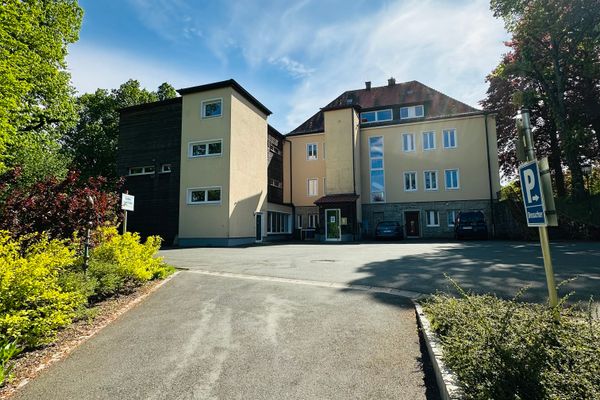 Das GER-Haus ist von hinten zu sehen, eingebettet in eine ruhige Umgebung mit vielen Bäumen. Direkt daneben befindet sich ein großer Parkplatz, der ausreichend Platz zum Parken bietet. Für Menschen mit eingeschränkter Mobilität steht ein Aufzug zur Verfügung, der einen barrierefreien Zugang zum Gebäude ermöglicht. So ist das Haus auch für Besucher mit Rollstuhl oder Kinderwagen bequem erreichbar.