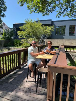 Auf dem Bild sind 2 Personen im Außenbereich der Bar zu sehen, die mit Bier anstoßend in die Kamera blicken.