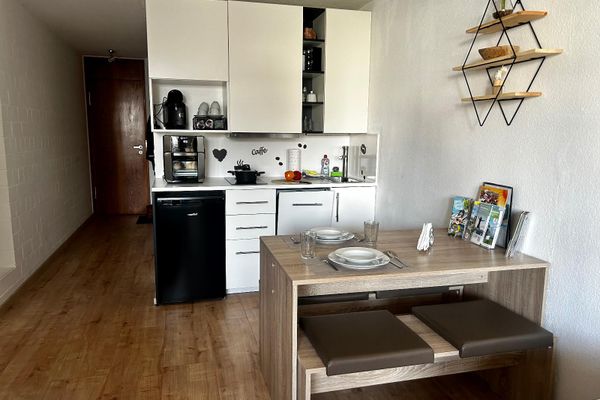 Eine kleine Küchenzeile mit Herd, Spülmaschine und Kaffeeautomat. Über dem Essbereich befindet sich ein Wandbord mit drei Brettern.