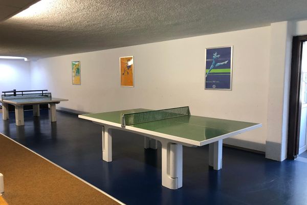 In einem Raum stehen hintereinander zwei Tischtennisplatten auf einem Kunststoffboden. Daneben verläuft ein Bereich mit Korkboden. An der Wand hängen drei Bilder mit Sportmotiven.