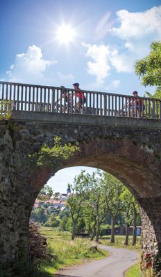 Eine Gruppe von vier Radfahrern fährt auf einer hohen, steinernen Brücke von rechts nach links. Durch den runden Brückenbogen sieht man eine geschlängelte Straße mit einer Baumreihe und an deren Ende einen Ort mit Kirchturm.