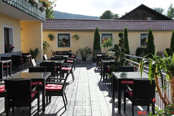 Auf der Terrasse stehen viereckige Tische mit passenden Rattanstühlen, dazwischen mehrere Grünpflanzen. Im Hintergrund begrenzt ein langgezogenes, teils mit Pflanzen und Blumen geschmücktes Gebäude die Terrasse.