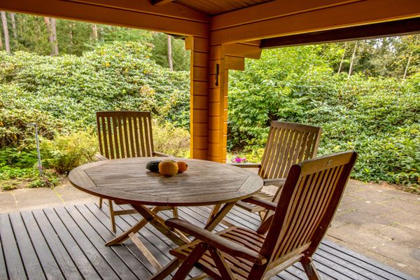 Überdachte Terrasse mit rundem Holztisch und drei Stühlen, Blick in einen dicht bepflanzten Garten.