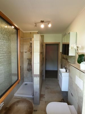 Ein modernes Badezimmer mit halboffener, begehbarer Dusche, WC, Waschbecken mit Unterschrank und Spiegel. Die Fliesen sind cremefarben gemustert.