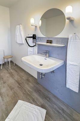 Das Bild zeigt ein modernes Badezimmer mit einem Waschbecken, darüber ein runder Spiegel und zwei Wandlampen, die den Raum erhellen. Links hängt ein Handtuch an einem Haken, rechts ein weiteres an einer Stange, und ein Haartrockner ist an der Wand befestigt.