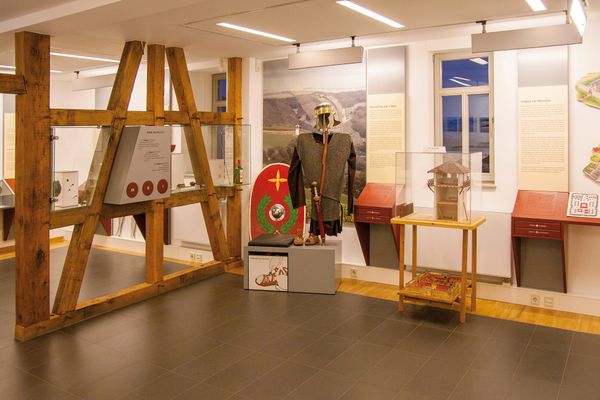 Blick in den Ausstellungsraum mit einer römischen Uniform, einem Schild links daneben und dem Modell eines Wachturms in einer Vitrine rechts daneben. Linker Hand teilt ein offenes Fachwerk den großen Raum. Im Fachwerk ist ebenfalls eine Ausstellungfläche mit Textinformationen. Weitere Tafeln mit Bildern und Inoformationen befinden sich an der Wand.