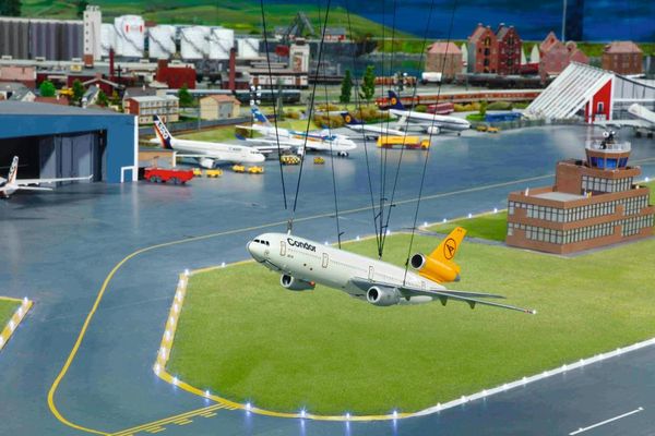 Mini-Flughafengelände mit Terminal, Hangar und einigen Flugzeugen am Boden. Ein Flieger befindet sich kurz nach dem Start in der Luft.