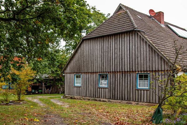 Haus mit Holzfassade, mehreren Fenstern und Garten mit Bäumen und gepflastertem Weg.