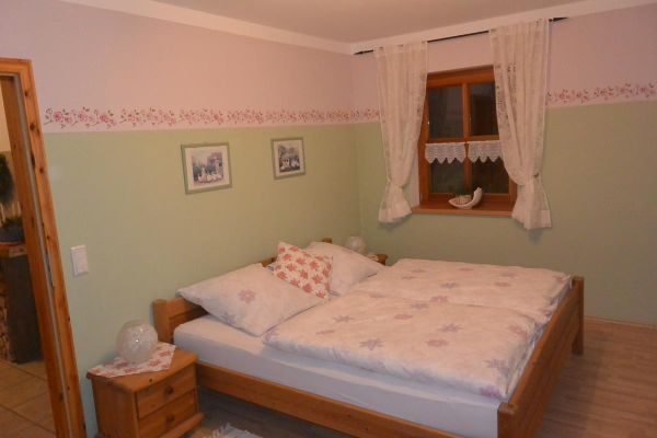 Ein Schlafzimmer mit einem Holz-Doppelbett, einem Nachttisch und einer luftigen Schalgardine vor dem Sprossenfenster. Die Wände sind in hellen und mintgrünen Farben gestrichen, unterteilt durch eine Blumenbordüre.