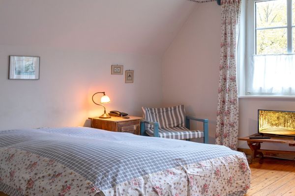 Schlafzimmer mit Bett, kleinem Tisch mit Fernseher, Stuhl und Fenster mit Vorhängen.