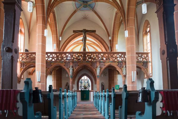 Blick auf den Chorraum der Kirche mit einer kunstvoll gefertigten Trennwand aus Bögen und Ballustraden aus Sandstein vor dem hölzernen Kreuz mit der Jesusstatue. Alle Säusen sind aus rotem Sandstein und alle spitz zulaufenden Bögen an den Kanten ebenfalls. Die blauen Bankreihen geben einen zentralen Mittelgang frei, der direkt auf den Altar zuführt, der mit einem grünen Ornament gedeckt ist. in einem Raum hinter dem Altar sind drei bunt bemalte Holzfiguren r zu sehen.