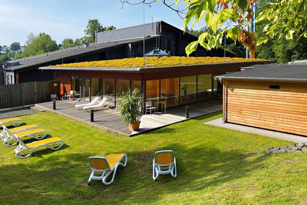 Auf der grünen Wiese stehen rund um eine hölzerne Terrasse eingie gelb-weiße Kunststoffliegen. Auf der Terrasse, die um die Ecke eines Falchdachbaus gebaut wurde, steht eine Palme im Topf und ein paar Stühle sowie zwei Liegen stehen bereit. Der Flachdachbach ist begrünt und die Seiten bestehen fast nur aus Fensterfronten, hinter denen man schemenhaft Saunainventar erkennt. Hinter dem Flachdachbau schließt sich ein höherer Bauabschnitt mit versetztem Pultdach an. Dahinter sind Baukronen und in der Ferne Häuser zu erkennen. Der Himmel ist klar und blau, der Ast eines Laubbaumes ragt von rechts oben in das Bild. Am rechten Rand steht noch ein kleiner Holzbau.