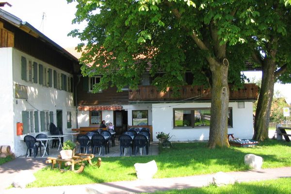 Der Betrachter blickt auf das Gasthaus und dem Bereich der Außengastronomie. Am Rande stehen zwei große Bäume.