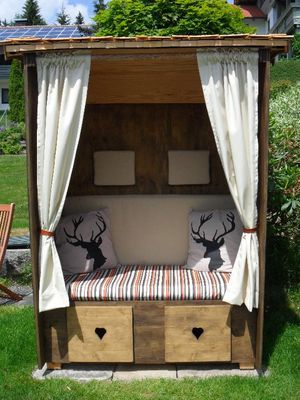 Im Garten steht ein Bayerwald-Strandkorb aus Holz mit zurückgezogenen, hellen Vorhängen und Kissen mit Hirschkopfmotiv.